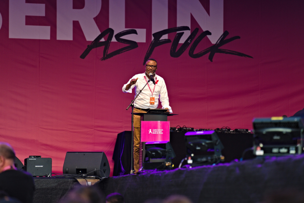 Ein Mann in einem weißen Hemd und braunen Hosen steht auf einer Bühne und hält ein Mikrofon in der Hand, mit einem pinken "Berlin as Fuck"-Plakat dahinter und einer Menge davor.