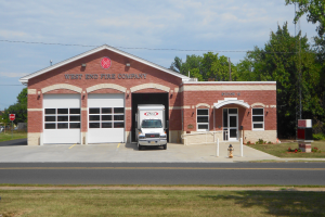 Eine Feuerwache mit einem vor ihr geparkten Löschfahrzeug, das Fenster, Türen und Text an der Wand hat, mit einem Feuerhydranten im Vordergrund und Bäumen, Strommasten und einem bewölkten Himmel im Hintergrund.