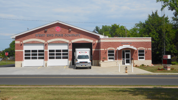 Eine Feuerwache mit einem vor ihr geparkten Löschfahrzeug, das Fenster, Türen und Text an der Wand hat, mit einem Feuerhydranten im Vordergrund und Bäumen, Strommasten und einem bewölkten Himmel im Hintergrund.
