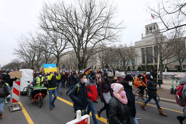 Eine große Gruppe von Menschen marschiert bei einer Protestaktion auf einer Straße in Washington, D.C., mit Schildern und Transparenten, einige fahren Fahrräder, unter einem klaren blauen Himmel.