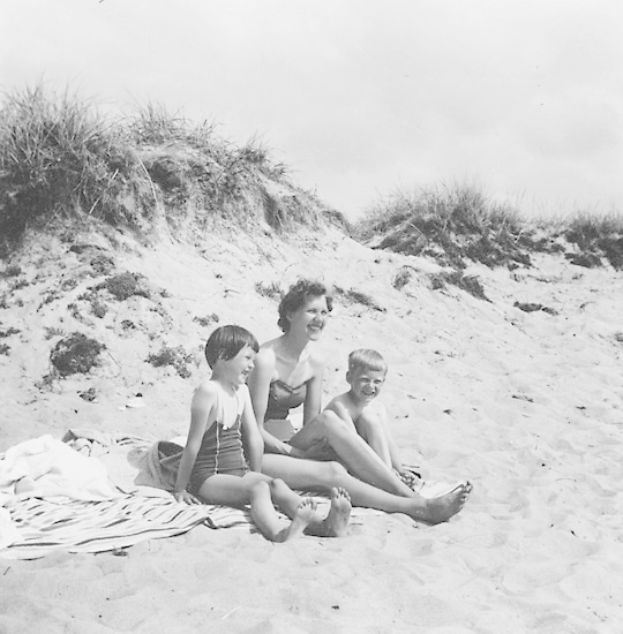 Schwarz-weiß-Foto einer Frau in einem Bikini und zwei Kindern in Badebekleidung, die auf einer Decke auf einem sandigen Strand mit Gras und Himmel im Hintergrund sitzen.