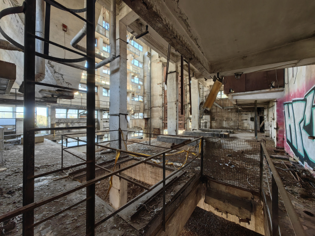 Verlassenes Gebäudeinnere mit Graffiti-beschmierten Wänden, bröckelnden Oberflächen, kaputten Fenstern, Geländern, Pfosten, Rohren und anderem Schutt, mit einem Wasserzeichen unten.