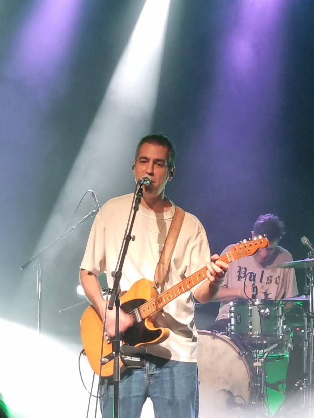 Ein Mann spielt eine E-Gitarre auf der Bühne, während er in ein Mikrofon singt, wobei ein Schlagzeuger hinter ihm sichtbar ist.