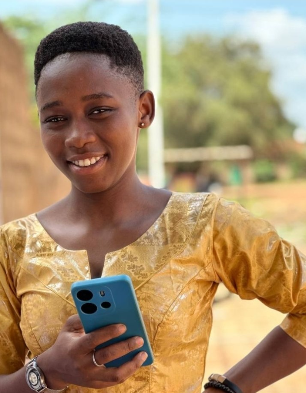 Ein junges Mädchen in einem gelben Kleid hält ein blaues Telefon, lächelt vor einem Hintergrund aus Wand, Pfahl, Bäumen und Himmel.