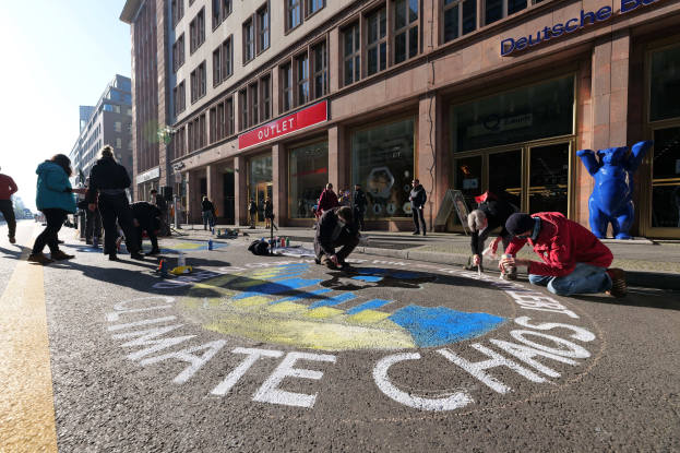 Eine Gruppe von Menschen sitzt auf dem Boden vor einem Gebäude während einer Klimawandel-Demonstration in Berlin, umgeben von Flaschen und anderen Gegenständen, mit Bäumen und einem klaren blauen Himmel im Hintergrund.