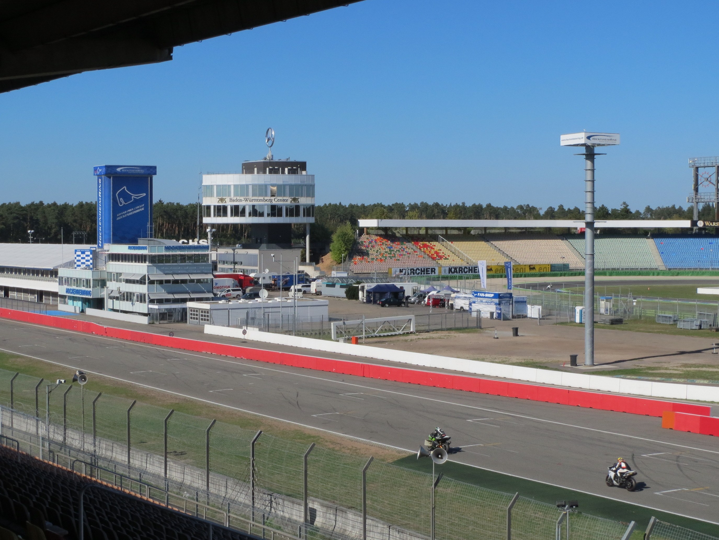 Gruppe von Motorradfahrern auf einer Rennstrecke, umgeben von einer Absperrung, Gras, Fahrzeugen, einem Gebäude, Schildern, Pfosten, einem Stadion, Lampen, Bäumen und einer bewölkten Himmel.
