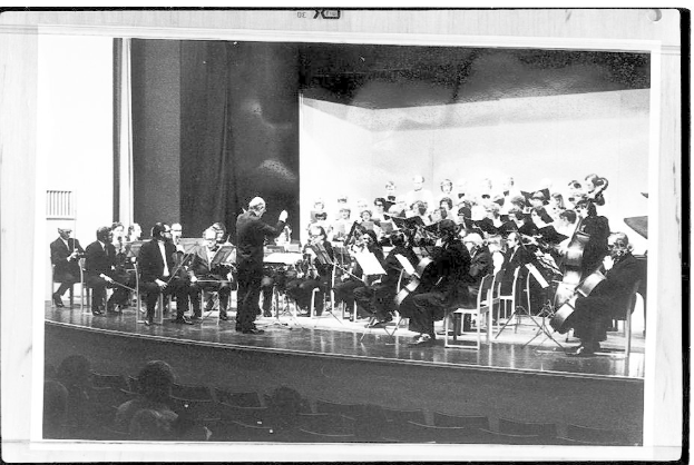 Ein Dirigent in Anzug und Krawatte steht auf einer Bühne und leitet ein Orchester mit Geigen, Celli und Flöten, während das Publikum auf Stühlen aufmerksam zuhört.