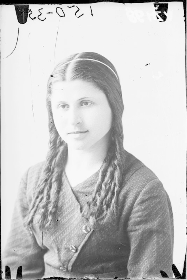 Ein Schwarz-Weiß-Porträt von Julia Margaret Cameron, eine Frau mit langen Zöpfen, die zu einem Dutt gebunden sind und ein weißes Kleid sowie einen gemusterten Schal trägt, mit einem sanften Gesichtsausdruck und ihren Händen vor der Brust verschränkt.