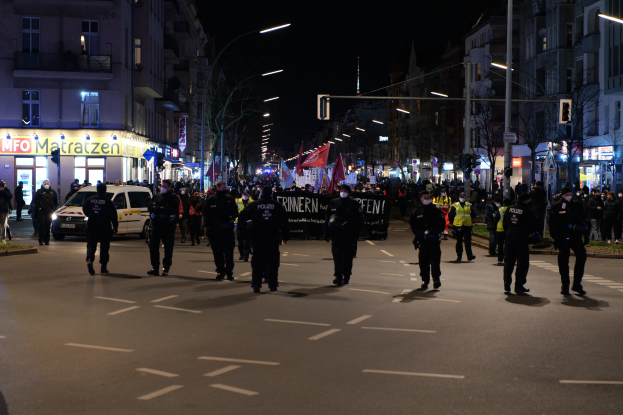 Eine Gruppe von Menschen geht ├Ąchtlich auf einer Stra├če in Berlin, Deutschland, mit Geb├Ąuden, B├Ąumen, Stra├čenlaternen, Verkehrsampeln, Fahrzeugen und einem Transparent im Hintergrund.