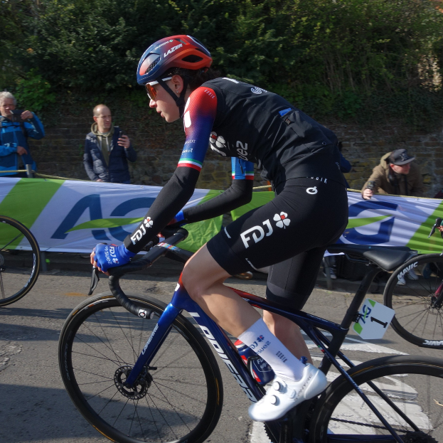 Professionelle Radrennfahrerin Katie Archibald fährt auf der Straße mit Helm, Brille, Handschuhen und Schuhen, umgeben von einer Menge und einem Banner.