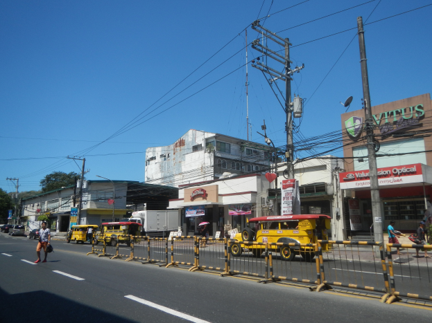 Eine belebte Stadtstraße mit starkem Verkehr, Fußgänger auf dem Gehweg, hohe Gebäude mit Beschriftung, Bäume und einen klaren blauen Himmel.