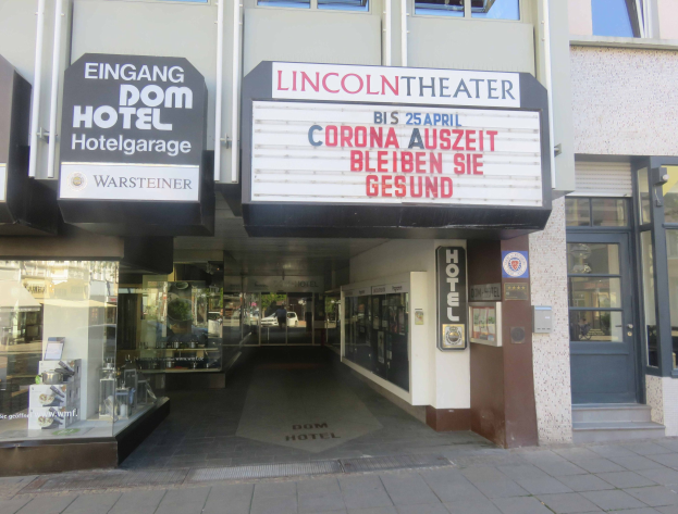 Außenansicht des Lincoln Theaters in Berlin, Deutschland, mit Glasfenstern, Türen und einer Texttafel sowie Innenraum-Elementen, die eine lebendige Stadtlandschaft suggerieren.