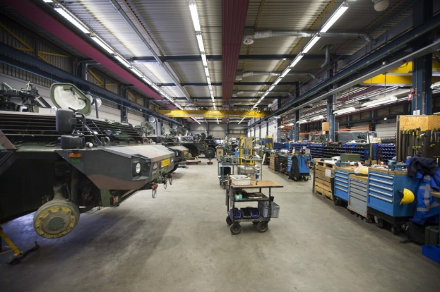 Großes Lager mit Maschinen, Fahrzeugen links, Wagen mit Gegenständen und Holzboxen rechts und Deckenbeleuchtung.