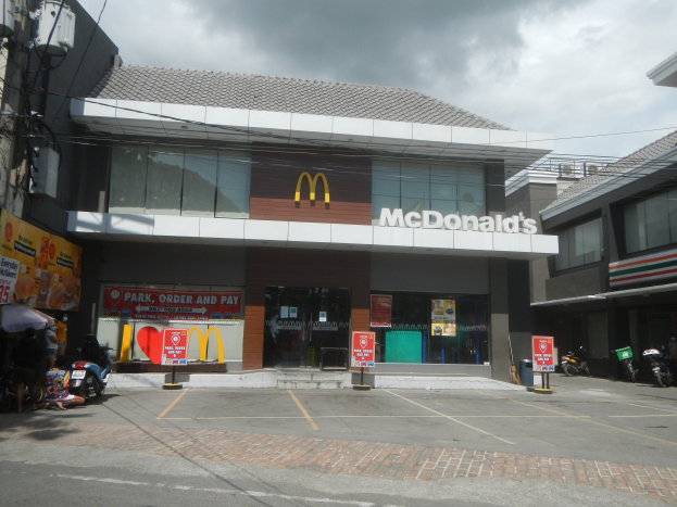 Ein McDonald's Restaurant an einer Straßenecke mit umliegenden Gebäuden, Motorrädern, Fußgängern, Regenschirmen, Werbetafeln, Strommasten, Kabeln und einem bewölkten Himmel.