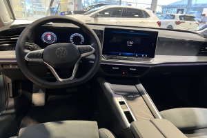 Das Innere eines Volkswagen e-Golf, mit einem Lenkrad, Armaturenbrett, Bildschirmen, Knöpfen, Sitzen und Glastüren, mit einem Blick auf andere Autos, Gebäude, Bäume, Pfosten und den Himmel durch die Glastüren.