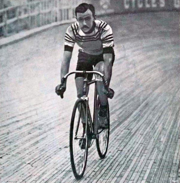 Schwarz-weißes Foto eines Mannes, der auf einer Bahn einen Fahrrad fährt, mit einem Zaun und einer Tafel im Hintergrund.