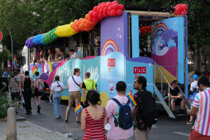 Eine Gruppe von Menschen, die eine Straße entlanggehen, neben einem Lastwagen mit bunten Luftballons, mit Schildern an den Straßenlaternen und Bäumen und Gebäuden im Hintergrund, was auf eine Pride-Parade in Paris hindeutet.
