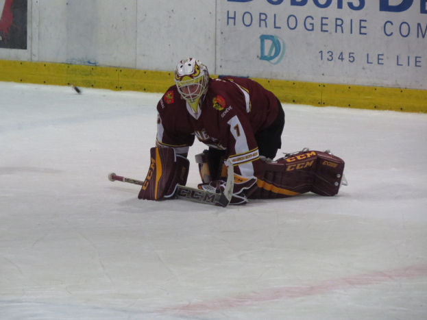 Eishockeyspieler in rot-gelber Uniform, der einen Schuss auf dem Eis abwehrt, trägt Helm, Handschuhe und Knieprotektoren und hält einen Eishockeyschläger, vor einer Wand mit Text im Hintergrund.