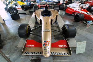 Rennwagen in einem Motorsportmuseum ausgestellt, getrennt durch einen Metallzaun mit Informationsschild im Vordergrund.