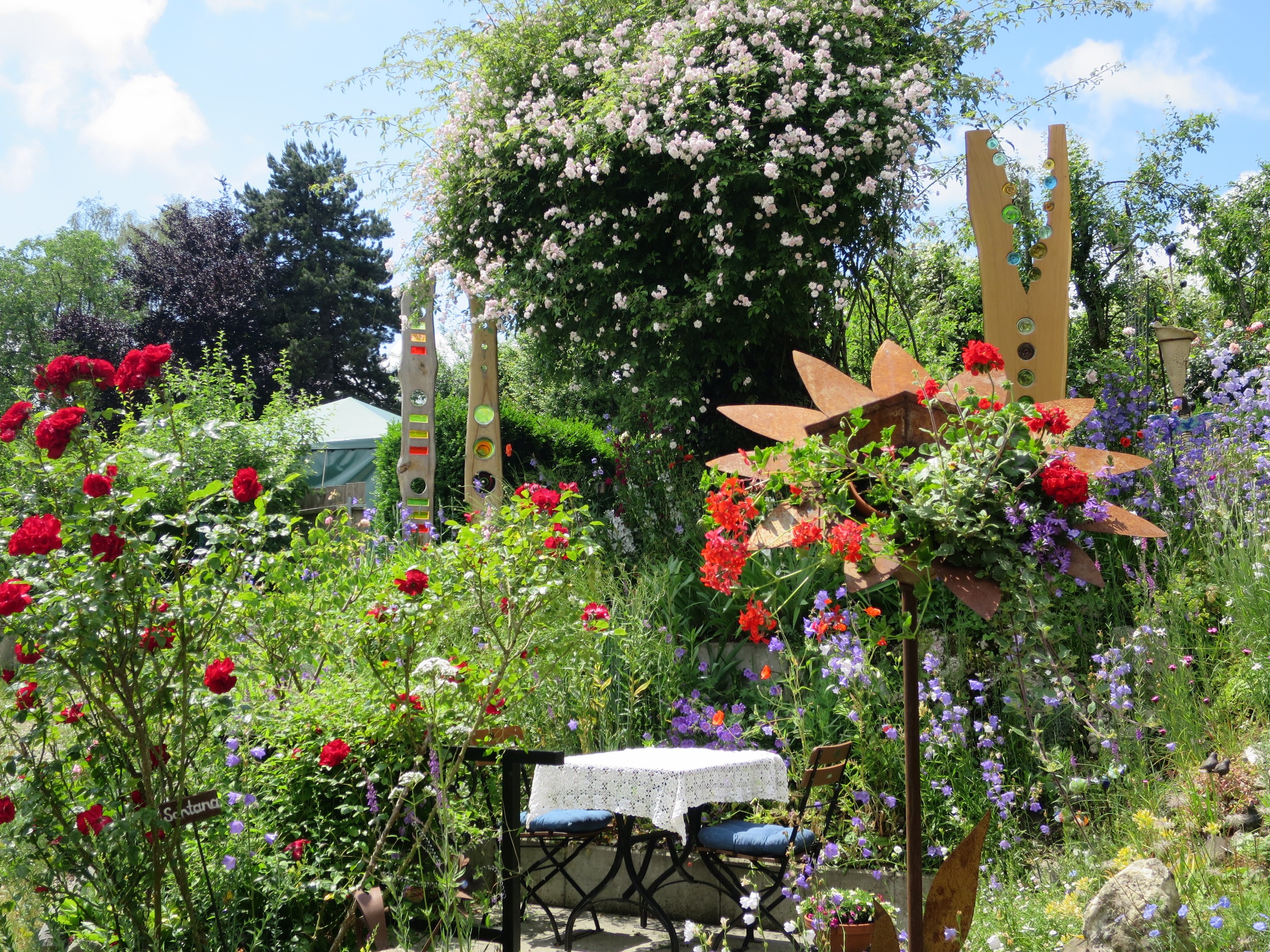 Ein Garten mit bunten Blumen und Pflanzen, der einen Tisch mit einer Decke und Stühlen zeigt, umgeben von Bäumen unter einem bewölkten Himmel auf dem Chelsea Flower Show.