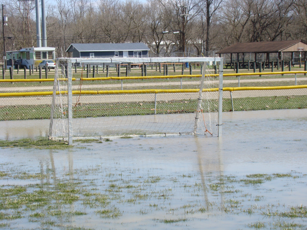 Fußballtor in einem überfluteten Feld mit Hütten, Pfählen, Bäumen, Fahrzeugen und einem klaren blauen Himmel im Hintergrund.
