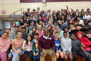 Gruppe von Schülern, die in einem Turnsaal für ein Foto posieren, einige sitzen auf der Treppe und andere stehen, eine Wand im Hintergrund, eine Tür und ein Ausgangsschild auf der linken Seite.