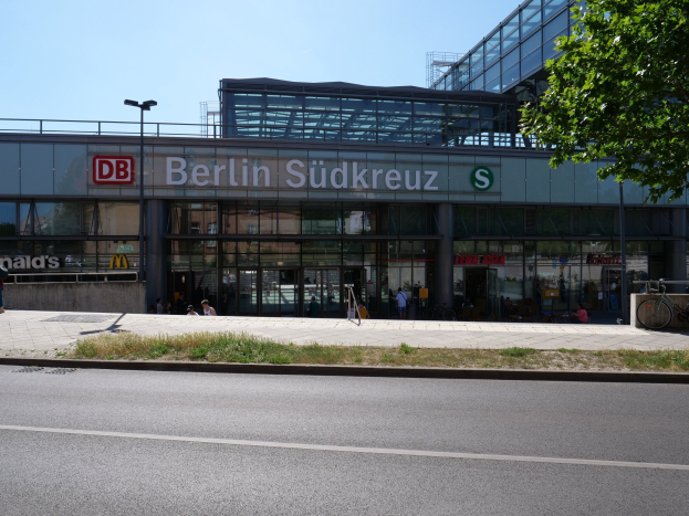 Ein großes Glaswand-Bahnhof mit der Aufschrift "Südkreuz" in Berlin, Deutschland, mit Straßenlaternen, Lampen, Fahrzeugen, Fußgängern, Fahrrädern, Bäumen und einem klaren blauen Himmel.