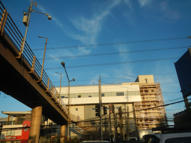 Eine Stadtstraße mit parkenden Autos, eine Brücke mit Geländern, Laternen, Strommasten mit Kabeln, Ampeln, Gebäude mit Fenstern und ein bewölkter Himmel im Hintergrund.