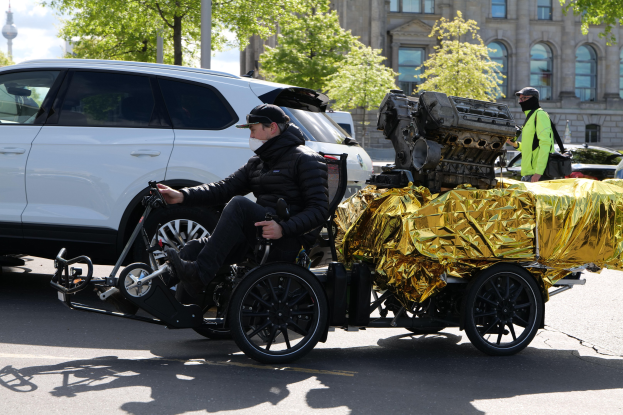 Ein Mann im Rollstuhl mit einem großen Motor auf dem Rücken, umgeben von Fahrzeugen auf der Straße, mit Bäumen, Gebäuden, Polen und einem klaren blauen Himmel im Hintergrund.