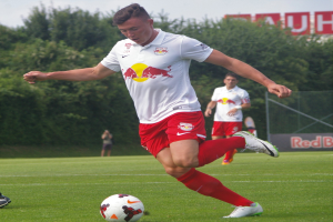 Ein Mann in einem Fußballtrikot kickt einen Ball auf einem grünen Feld mit Bäumen und einem klaren blauen Himmel im Hintergrund, mit einem Schild mit der Aufschrift "RB Leipzig"