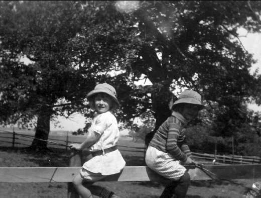 Zwei junge Jungs in Mützen halten Baseballschläger in einem grünen Park mit Bäumen und einem Zaun im Hintergrund, aufgenommen in Schwarz-Weiß.