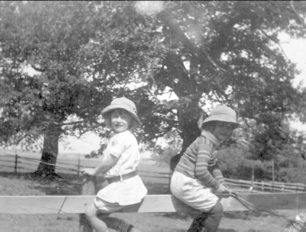 Zwei junge Jungs in Mützen halten Baseballschläger in einem grünen Park mit Bäumen und einem Zaun im Hintergrund, aufgenommen in Schwarz-Weiß.