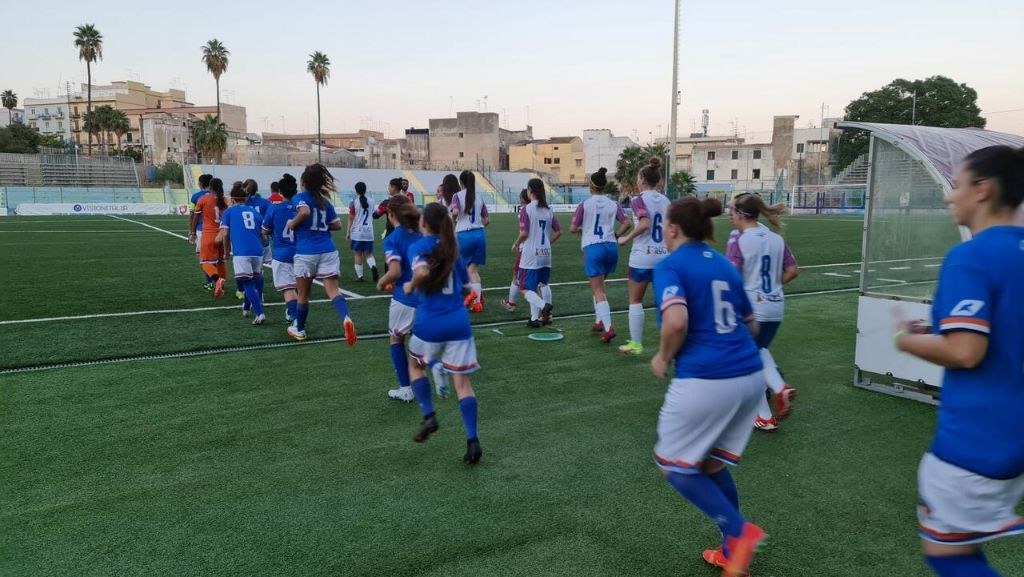 Eine Gruppe von Frauen, die auf einem Outdoor-Feld Fußball spielen, mit Gebäuden, Bäumen und einem Torpfosten im Hintergrund.