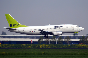 Ein AirBaltic Airbus A320-200 startet vom Frankfurter Flughafen, umgeben von grünem Gras und bunten Blumen, mit Gebäuden, Bäumen, Pfählen und Fahrzeugen im Hintergrund.