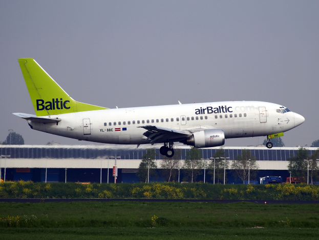 Ein AirBaltic Airbus A320-200 startet vom Frankfurter Flughafen, umgeben von grünem Gras und bunten Blumen, mit Gebäuden, Bäumen, Pfählen und Fahrzeugen im Hintergrund.