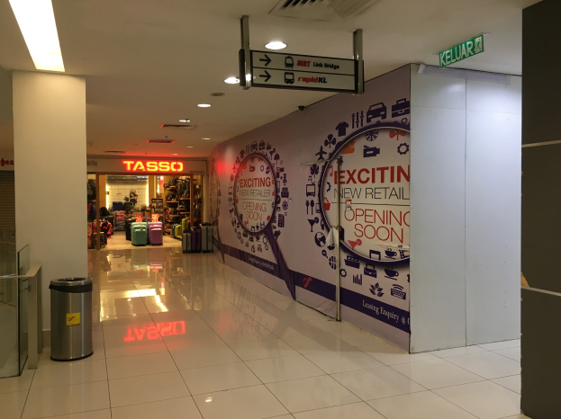 Entrance to a shopping mall with a "Exciting New Retail Opening Soon" sign, a dustbin on the left, a glass door on the right, and ceiling lights above.