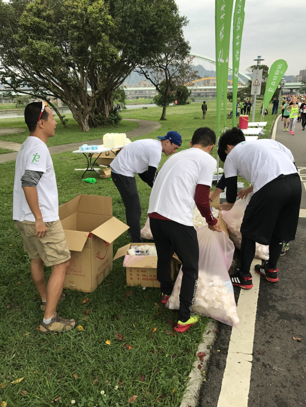 Gruppe von Menschen an einer Straße mit verstreuten Kartonboxen, Flaschen und Schildern, die an einem Spendenlauf teilnehmen mit Bäumen, Gebäuden und einem klaren blauen Himmel im Hintergrund.
