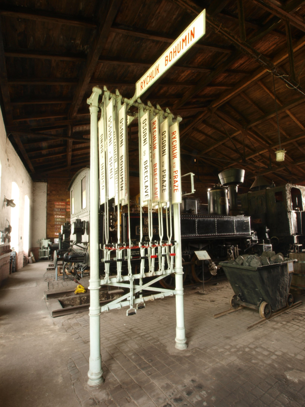 Ein Bahnsteig mit einem Zug auf den Gleisen, einem Bodenwagen, einem Schild, einer Wand links, einer Decke darüber und einer zentralen Signalbox.