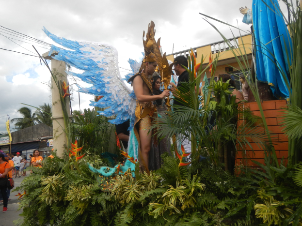 Eine Gruppe von Menschen geht eine Straße entlang neben einem blumengeschmückten Faschingswagen mit kostümierten Personen, mit Gebäuden, Bäumen, Strommasten und einem bewölkten Himmel im Hintergrund.