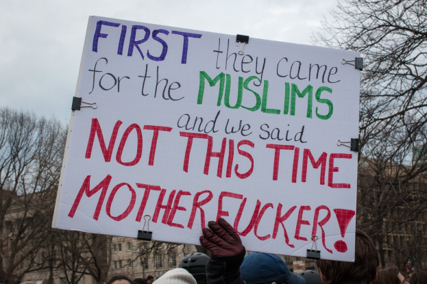 Eine Gruppe von Menschen mit Mützen und Handschuhen hält ein Schild mit der Aufschrift "Zuerst kamen sie wegen der Muslime und wir sagten nicht dieses Mal, verdammt" mit Bäumen, Gebäuden und einem klaren blauen Himmel im Hintergrund.