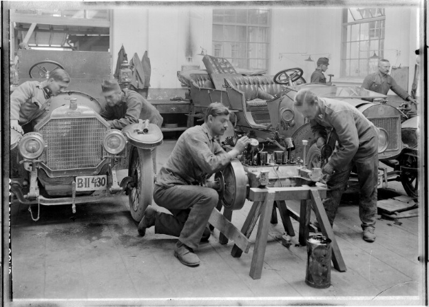 Ein Schwarz-Weiß-Bild von mehreren uniformierten Männern, die an einem Auto in einer unordentlichen Garage arbeiten, umgeben von Werkzeugen und Equipment.