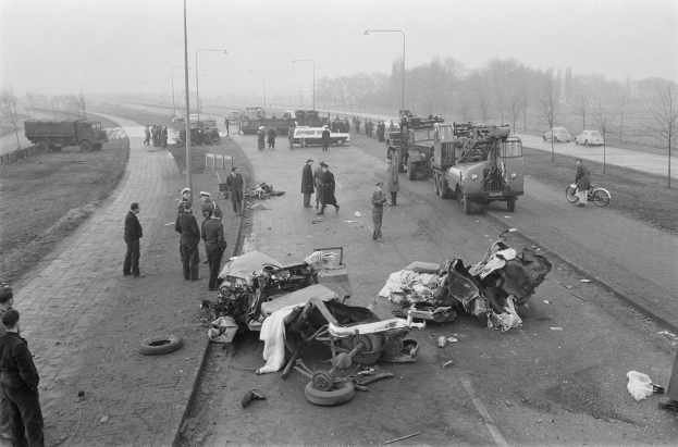Schwarz-weiß-Szene eines Autounfalls am Straßenrand mit mehreren Fahrzeugen, darunter ein Landfahrzeug, und einer Gruppe von Menschen, die darum herumstehen, mit Laternenpfählen, Bäumen und Himmel im Hintergrund.