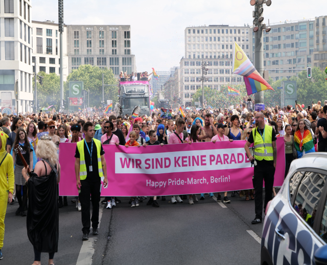 Eine große Gruppe von Menschen bei der 'Happy Pride March Berlin' mit einem Banner, daneben ein geparktes Auto, Gebäude und Bäume im Hintergrund unter einem klaren blauen Himmel.