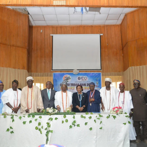 Gruppe von Menschen um einen weißgedeckten Tisch mit Blumen und Blättern, im Hintergrund ein "ICCI International Trade Fair 2019"-Banner, Projektionsschirm und Deckenbeleuchtung.
