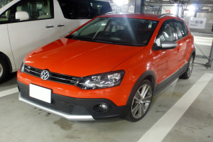 Orange Volkswagen Polo Cross 1.6 TDI Highline DSG neben einem weißen Lieferwagen in einer Tiefgarage mit Metallzaun, Texttafel, Lampen und Deckenlampen im Hintergrund geparkt.