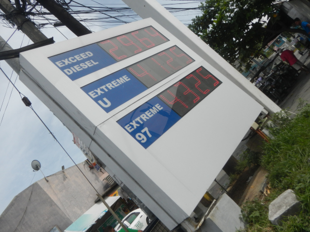 Eine Tankstelle mit einem Schild, auf dem "Exceed Diesel" steht, im Vordergrund, mit Strommasten, Gebäuden, Fahrzeugen, Menschen, Bäumen und anderen Objekten im Hintergrund unter einem sichtbaren Himmel.