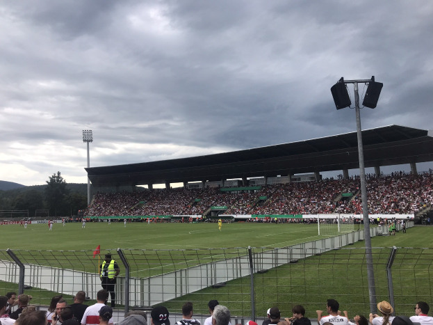 Große Zuschauermenge bei einem Fußballspiel in einem bewölkten, offenen Stadion mit beleuchteten Masten, umgeben von einem Zaun, Bäumen und Hügeln.