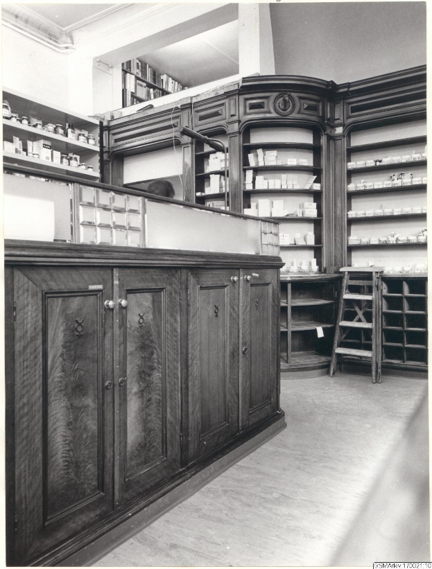 Schwarzes und wei├čes Foto eines vintage Apothekeninneren mit einer Theke, Schüben, einer Leiter und Regalen, die mit verschiedenen Gegenst├Ąnden bestückt sind, mit Text über dem Bild.