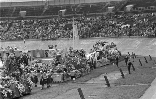 Schwarzes und weißes Foto eines Umzugs in einem Stadion mit stehenden und sitzenden Menschen, einem zentralen Springbrunnen und Blumenbouquets auf Fahrzeugen.