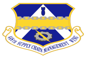 Ein Schild mit einem blauen Hintergrund, einem weißen Stern in der Mitte und einem weißen Banner mit der Aufschrift 'Lieferkette' in blau, umgeben von einem weißen Rand, mit der schwarzen, fetten Schrift '48th Supply Chain Management Wing' darüber und darunter.